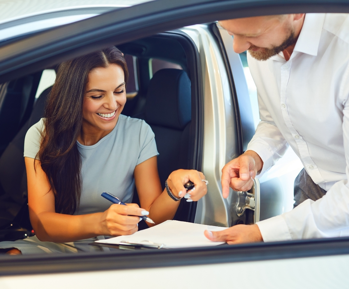 Car buyer signing paperwork while receiving keys from a dealership representative